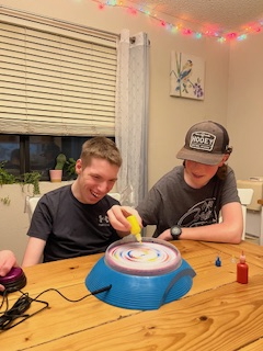 Two young men sitting at a table together with one places paint on the spinner while the other activates the spinner using a large button switch. 