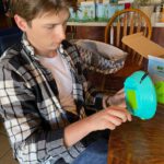 Young man with dark hair wearing a black and white checked shirt using tools to adapt a blue and green toy.