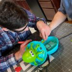 Man with dark hair and blue plaid shirt using tools to adapt a green and blue octopus toy.