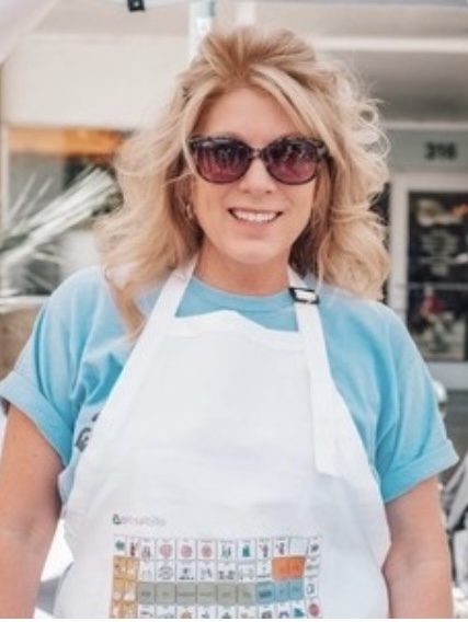 Blonde woman smiling wearing sunglasses, blue shirt, and apron