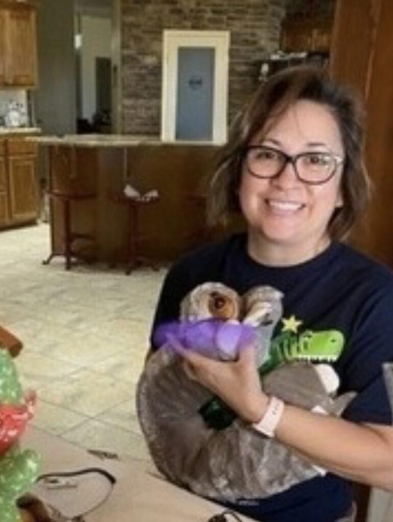 Dark haired woman smiling wearing glasses and dark t-shirt; holding adapted toys.